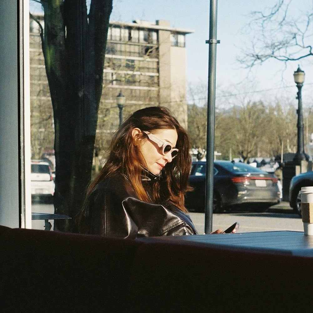 Mulher olhando o celular num café, com a rua da cidade ao fundo